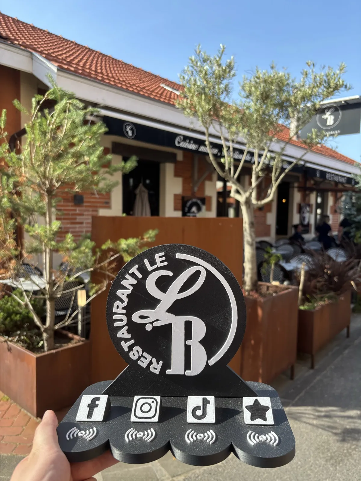 Stand NFC Filon Médoc du restaurant Le LB à Soulac-sur-Mer, posé en main devant la terrasse, avec ses 4 logos réseaux Facebook, Instagram, TikTok et avis Google